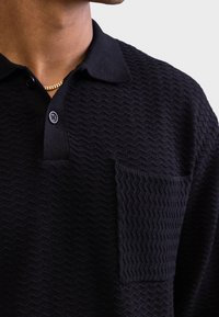 Polo preta com padrão texturizado em zig-zag, pala com botões e um bolso de aplicação único. Tecido macio com design clássico de gola.