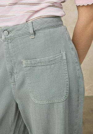 Nahaufnahme einer Person, die hellgrüne Jeanshosen mit einer großen Vordertasche und ein rosa gestreiftes Oberteil trägt.