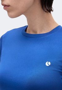 Άτομο που φοράει μπλε μπλουζάκι με στρογγυλό μικρό λευκό λογότυπο στο στήθος, απεικονίζεται από το λαιμό έως το πάνω μέρος του κορμού.