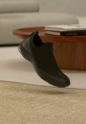 Chaussure noire sans lacets avec design à bout fendu flottant au-dessus d'un tapis beige, table ronde en bois et livres flous en arrière-plan.