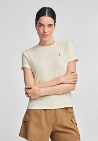 T-shirt in cotone beige chiaro con maniche corte, scollatura rotonda e un piccolo design ricamato sul petto. Tessuto liscio.