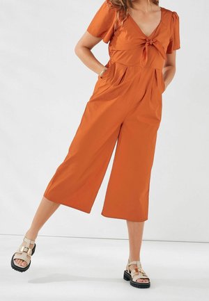 Jumpsuit i rustfarve med V-hals, knude foran, korte ærmer og brede, cropped ben. Styles med beige og sorte plateau-sandaler.