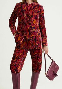 Costume en velours avec motif floral dans des tons de bordeaux, orange et jaune ; comprend une veste ajustée avec poches et un pantalon cintré. Sac assorti.