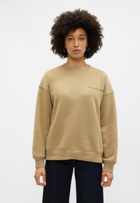 Kahovéná mikina s kulatým výstřihem a spadlými rameny, zdobená vyšitým textem na přední straně. V kombinaci s tmavě modrými džíny.