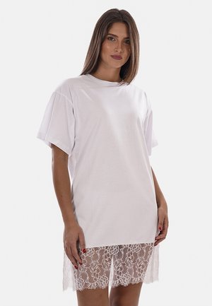 Donna che indossa un vestito T-shirt bianco oversize con maniche corte e un orlo in pizzo floreale trasparente, in piedi davanti a uno sfondo semplice.