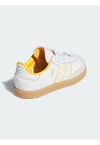 Weiße Leder-Sneaker mit gelben Akzenten an Ferse und Zunge, ausgestattet mit drei gelben Streifen und einer Gummisohle.