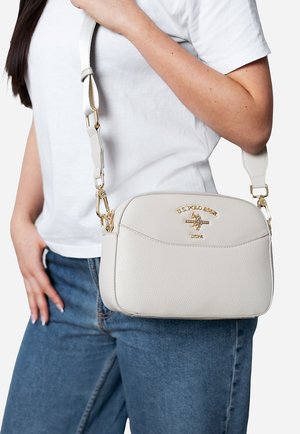 Frau trägt ein weißes T-Shirt und blaue Jeans und hält eine weiße Umhängetasche von U.S. Polo Assn. mit goldfarbenen Beschlägen.