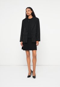 Blazer nero con decorazioni, indossato sopra un top e una gonna neri. L'outfit presenta rever affilati e un design su misura.