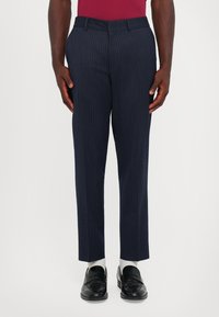 Uomo che indossa pantaloni cropped a righe blu scuro, calze bianche e mocassini in pelle nera, in piedi contro uno sfondo uniforme.