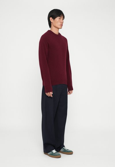 Maglione color bordeaux con scollo a giro, con maniche lunghe e polsini a coste, abbinato a pantaloni a righe navy e sneakers con dettagli verdi.
