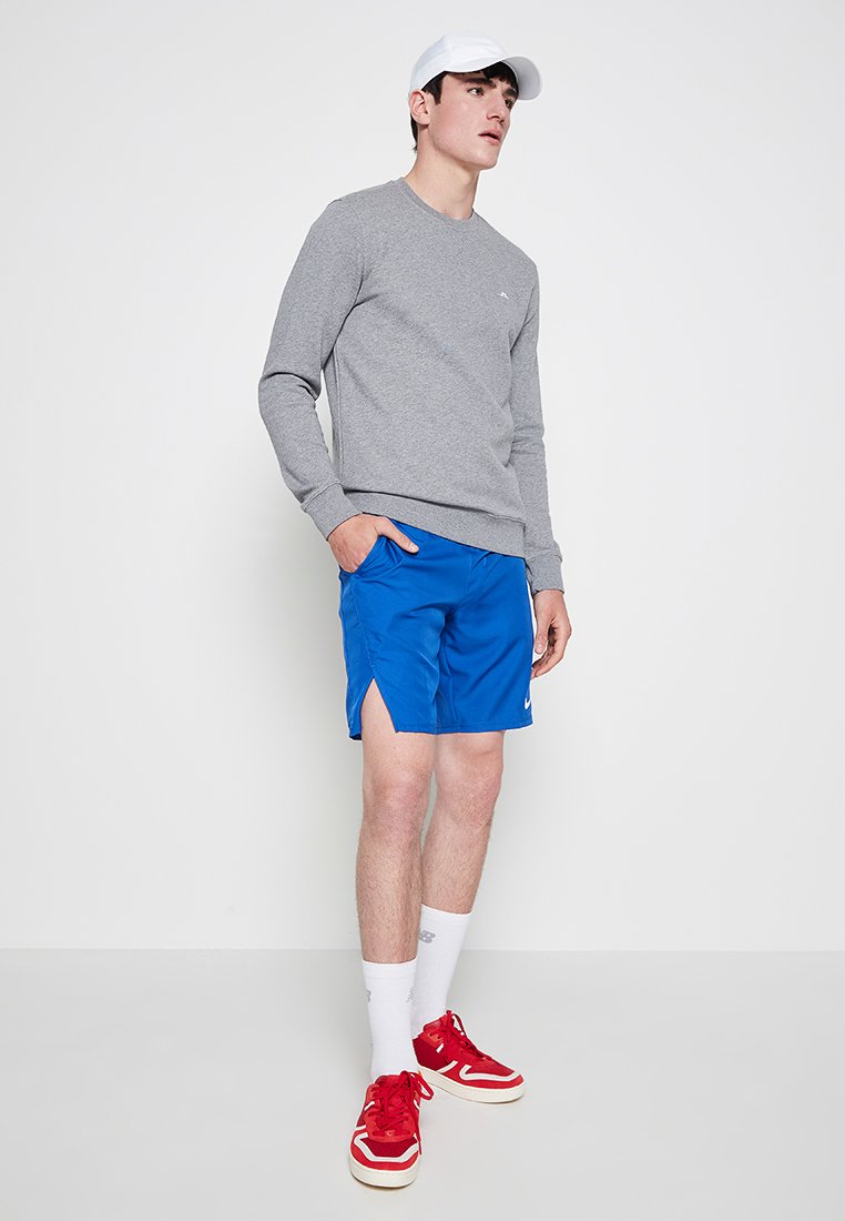 Grå tröja, blå träningsshorts, vit keps, röda sneakers och vita strumpor. Enkelt mönster, materialet verkar mjukt. Shorts med sidoflister.
