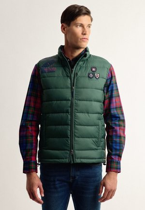 Chaleco acolchado verde con cierre de cremallera, con logotipos en el pecho. Llevado sobre una camisa de cuadros multicolor con tonos rojos, azules y verdes.