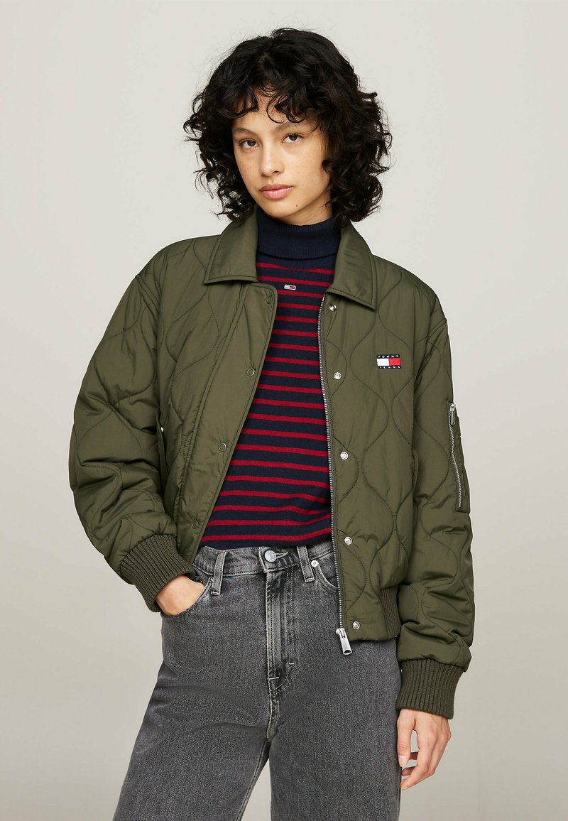 Chaqueta bomber acolchada de color verde oliva con puños y cuello de canalé, cierre de cremallera y un logo de parche. Lleva puesta sobre una camiseta a rayas en tonos marino y rojo y unos vaqueros grises.