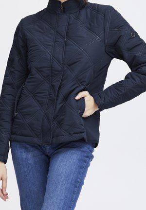 Persona con chaqueta acolchada azul marino con cremallera y bolsillos con botones a presión, combinada con vaqueros azules, mano en el bolsillo.