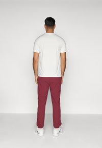 Balta īsām piedurknēm krekls ar tekstūras rakstu, apvienots ar bordo biksēm un baltām kedām. Skats rāda personu no aizmugures.