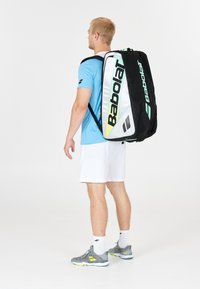 Mand i sportstøj bærer en stor Babolat tennis taske på ryggen, står op ad en ensfarvet hvid baggrund.