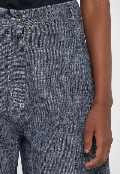 Pantaloni in denim caratterizzati da una trama testurizzata di colore blu scuro, con bottone frontale e zip, e dettagli di cucitura visibili. Mano parzialmente visibile.