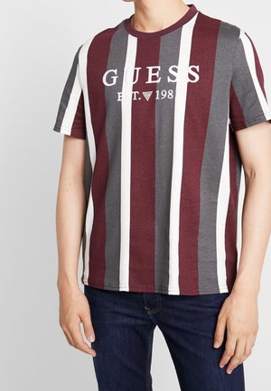 Homme portant une chemise à manches courtes avec des rayures verticales bordeaux, grises et blanches, arborant le logo "GUESS EST. 1981", associée à un jean bleu foncé.