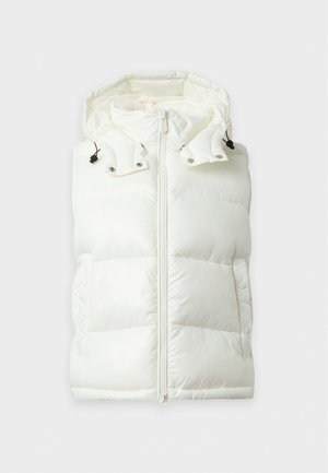 Femme portant une veste sans manches matelassée blanche, un haut à manches longues foncé, un pantalon large gris et des chaussures noires, debout devant un fond blanc.