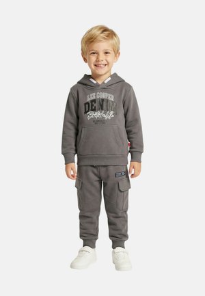 Garçon souriant portant un sweat à capuche gris foncé avec le texte "Lee Cooper Denim British UK" et un pantalon cargo assorti, debout en baskets blanches sur fond blanc.
