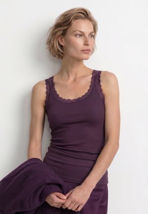 Femme blonde portant un débardeur violet bordé de dentelle, tenant un vêtement violet, regardant sur le côté sur fond blanc.