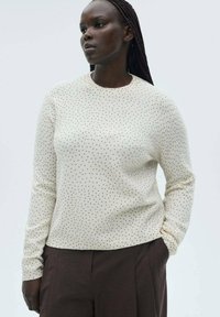Cremiger Pullover mit langen Ärmeln und schwarzen Punkten, runder Ausschnitt und lockerer Passform. Weicher Stoff, kombiniert mit braunen, hoch taillierten Hosen.