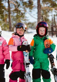 Dvě děti v barevném lyžařském oblečení: jedno v růžové s oranžovými akcenty, druhé v zelené. Obě drží lyže a mají přilby a rukavice.