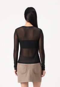 Haut transparent noir à manches longues avec une texture lisse et un col rond, porté avec une jupe taupe dotée de deux poches arrière.
