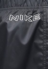 Μαύρο υφαντό νάιλον με ελαστική μέση, που διαθέτει ευδιάκριτο λευκό κεντημένο λογότυπο "NIKE" στο μπροστινό μέρος. Ομαλή υφή, μινιμαλιστικός σχεδιασμός.