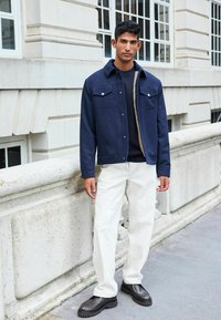 Navy blue jacket with button pockets over a textured navy sweater, paired with white trousers and brown chunky sole shoes. Neutral background.
