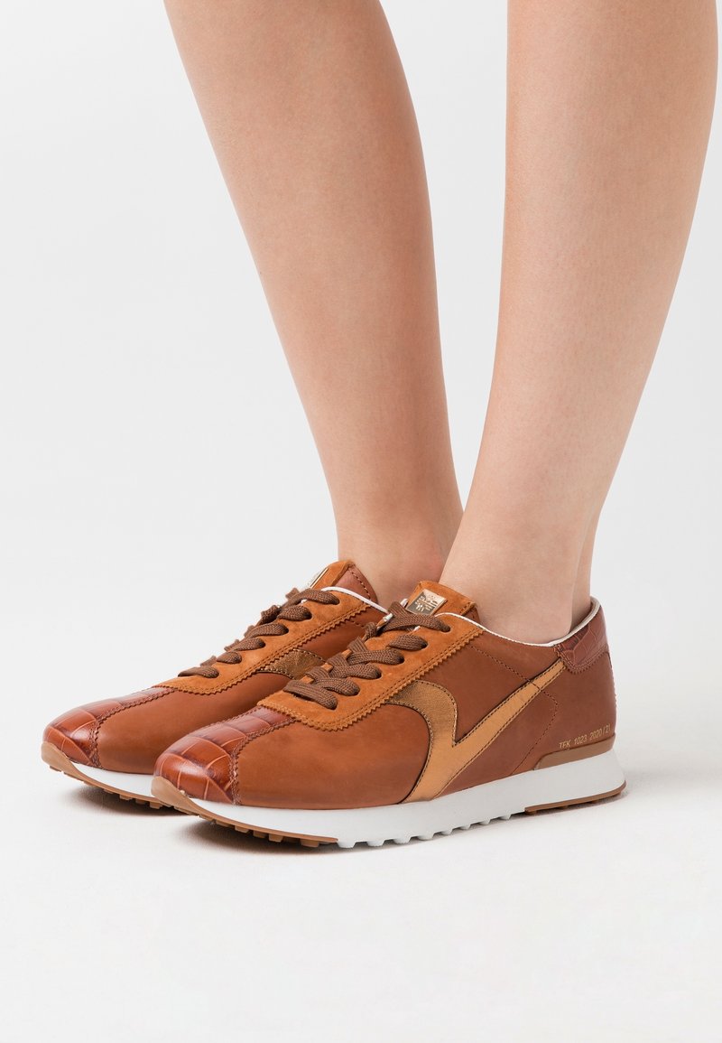 Baskets en cuir marron avec des semelles blanches portées pieds nus, montrant les détails latéraux et avant sur un fond clair uni.