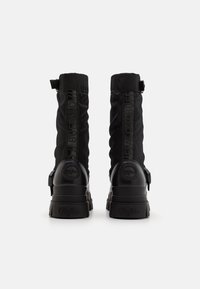 Bottes noires avec un haut en textile flasque, semelles en caoutchouc texturé et accents de logo à l'arrière, avec un détail de sangle.