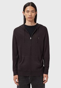 Ung man med långt mörkt hår iförd en svart zip-up huvtröja över en svart t-shirt, stående mot en enkel grå bakgrund.