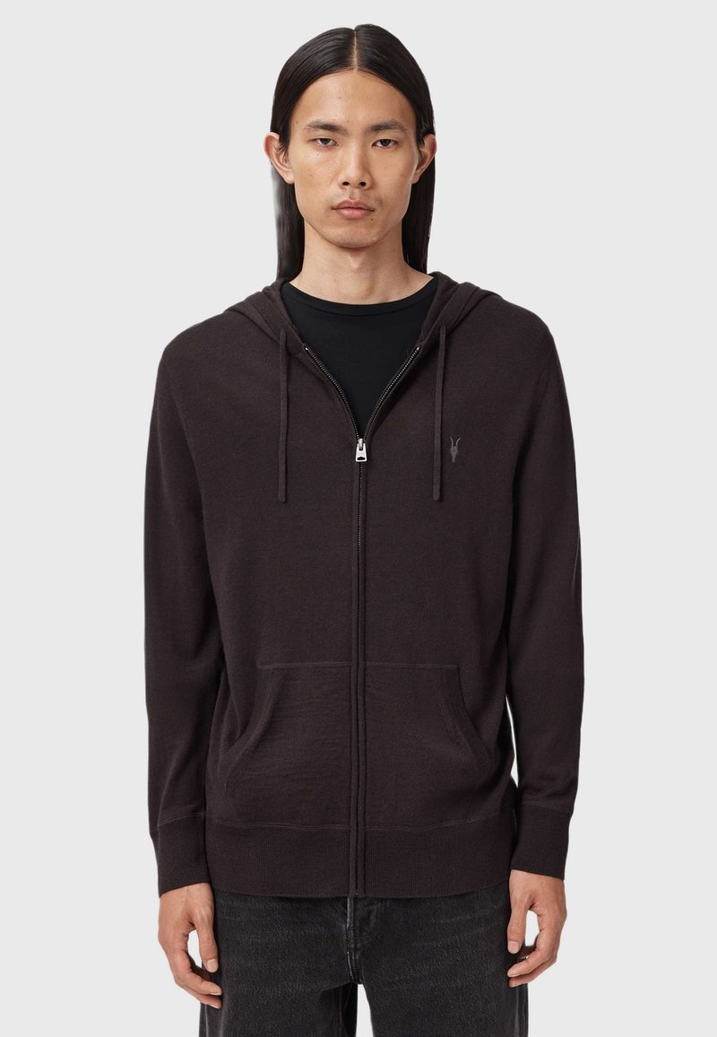Ung man med långt mörkt hår iförd en svart zip-up huvtröja över en svart t-shirt, stående mot en enkel grå bakgrund.