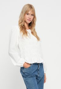 Hvid bluse med smock foran, puffede lange ærmer og flæsekanter. Båret med højthalsede blå denim jeans.