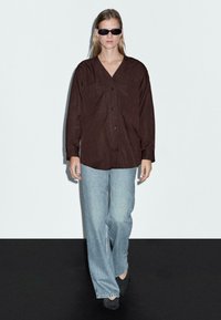 Person med sorte solbriller, mørkebrun knap-opp skjorte, lyseblå vide jeans og sorte spidse sko, der går på et sort og hvidt gulv.