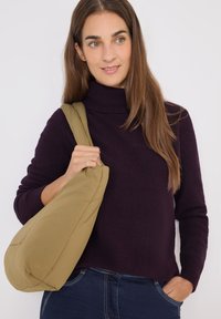 Beige Schultertasche mit runder Form, aus einem glatten, weichen Material, getragen von einer Person in einem dunkel-lila Rippensweater.