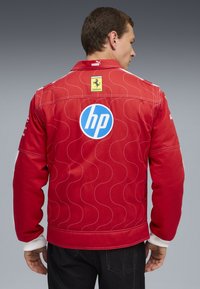 Uomo che indossa una giacca da corsa rossa Ferrari con cuciture bianche, con i loghi HP e Ferrari sulla schiena, visto da dietro su uno sfondo grigio.