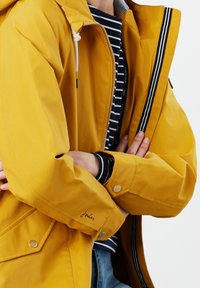 Żółta wodoodporna kurtka z kapturem, zapinana na zamek błyskawiczny, z paskami dekoracyjnymi. Posiada luźny krój, boczne kieszenie oraz detal z logo na mankiecie.