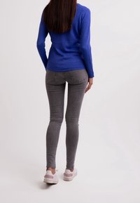 Vrouw staat met haar rug naar de camera, gekleed in een blauwe langemouwenshirt, grijze skinny jeans en witte sneakers met roze accenten.