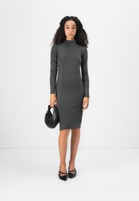 Tmavošedé priliehavé šaty s dlhým rukávom a vysokým golierom, s detailným prešívaním. Skombinované s čiernou kabelkou a topánkami s špicatou špičkou.