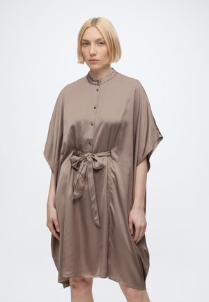 Kvinde med kort blondt hår iført en taupe satin knap-op kjole med brede ærmer og et bundet bælte i taljen, stående mod en ensfarvet hvid baggrund.