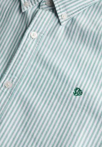 Gros plan d'une chemise à rayures verticales vertes et blanches, avec des boutons blancs et un petit logo vert brodé.