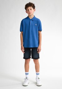 Modrá polos tričko s malým logem, tmavé denimové šortky, bílé tenisky a vzorované ponožky s modrými pruhy. Jednoduchý a ležérní design.