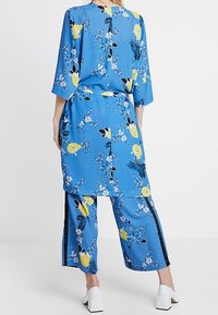 Modrý vzorovaný outfit s květinovým motivem a žlutými akcenty, volná tunika, kalhoty se širokými nohavicemi a černými bočními pruhy.