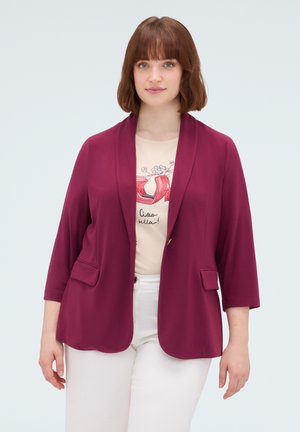 Kvinde med kort brunt hår iført en magenta blazer, beige grafisk t-shirt og hvide bukser, stående mod en ensfarvet lys baggrund.
