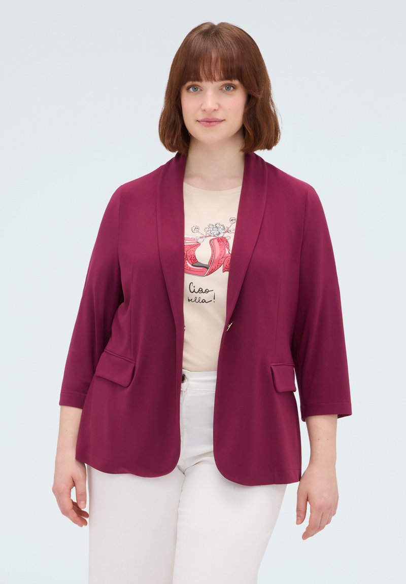 Frau mit kurzen braunen Haaren trägt einen magentafarbenen Blazer, ein beiges Grafik-T-Shirt und weiße Hose, steht vor einem schlichten hellen Hintergrund.