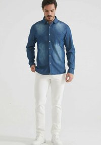 Chemise en jean bleu foncé avec un effet délavé, manches longues, poche poitrine ; portée avec un pantalon ajusté blanc et des baskets blanches.