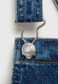 Denim jumpsuit har ett blått tyg med kontrasterande orange sömmar. En metallklämma fäster remmen och visar en varumärkeslogotyp.