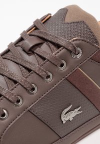 Brun sneaker med en blandning av texturerad läder och tyg, metallöglor och en logotyp som pryder. Snörena är också bruna med en subtil glans.